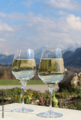 Zwei Gläser mit Weißwein. Im Hintergrund die Alpen. In den Gläsern spiegelt sich die Natur. Allgäu, Bayern, im Frühling