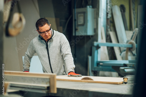 A craftsman in protective workwear is cutting wooden material on a saw in his workshop.