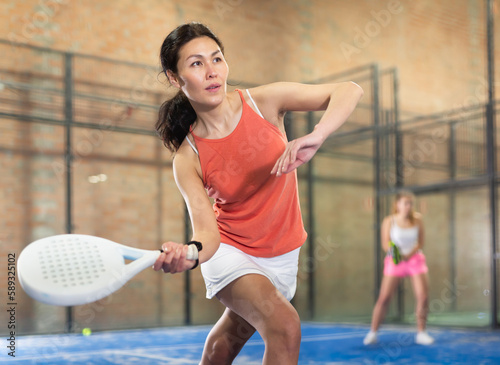 Positive asian female player hits the ball while playing padel at fitness health club © JackF