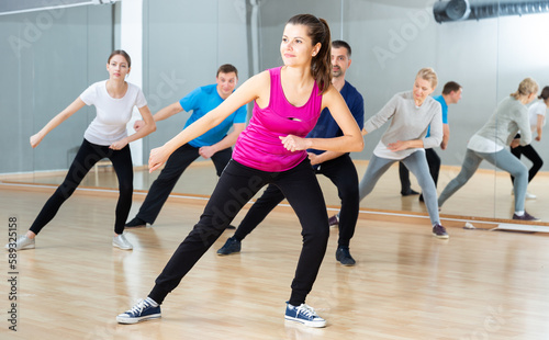 Positive young woman trainer doing aerobics exercises with group of adult people in dance center