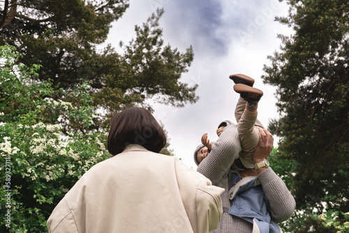 Low angle photo of multicultural family playing with baby, lifting, throwing infant up in air in nature. Unrecognizable father is holding kid in arms, while mother is watching at them. photo