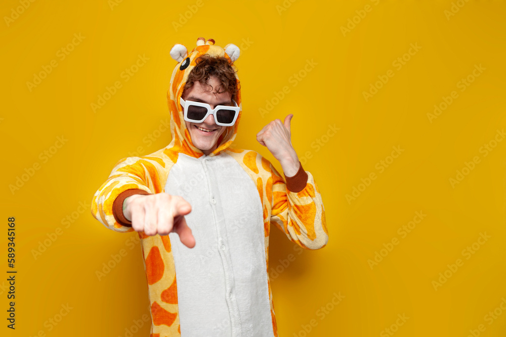 young joyful guy in funny children's giraffe pajamas and glasses invites you to party on yellow background