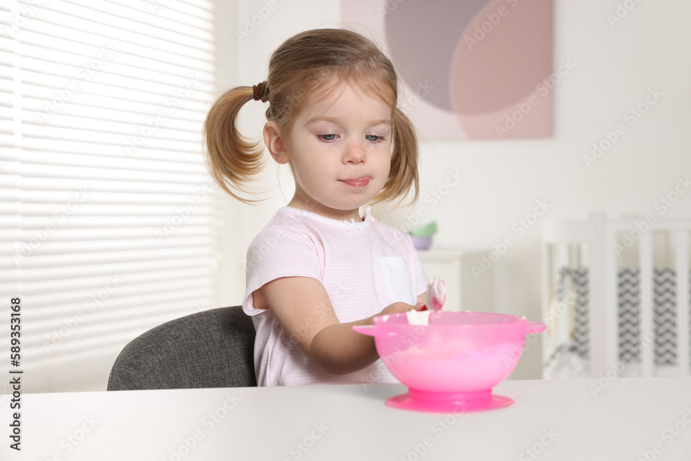 Cute little child eating tasty yogurt from plastic bowl with spoon at white table indoors Cute little child eating tasty yogurt from plastic bowl with spoon at white table indoors