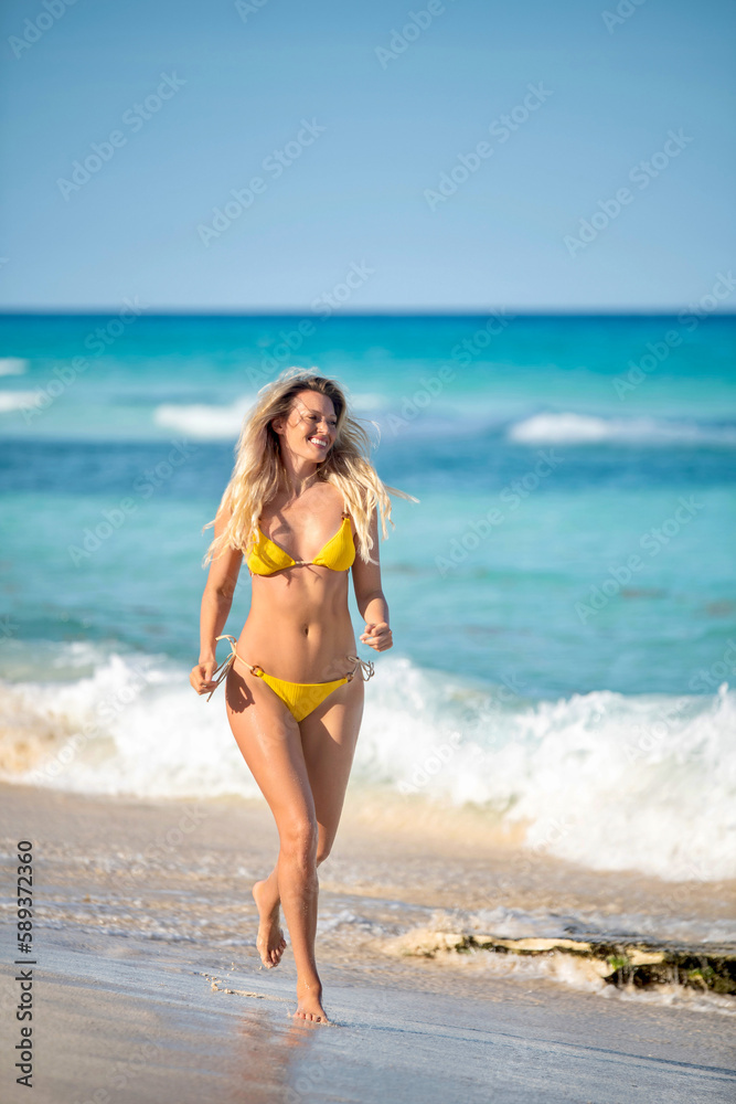Young blonde woman in a yellow bikini running on the beach enjoying the sun smiling