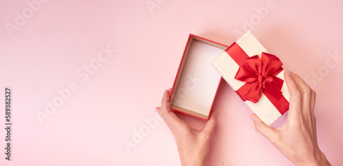 Hands holding open gift box with red bow on pink background. Top view