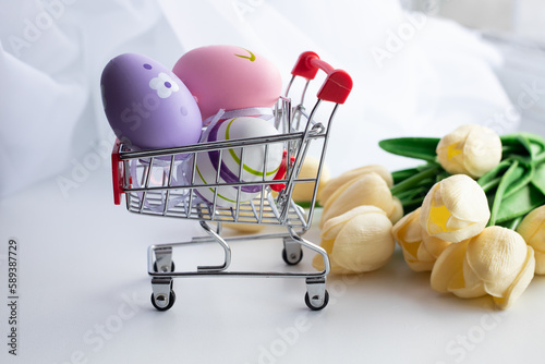 Colored eggs in a shopping cart on a white background with flowers. Easter Holiday Concept
