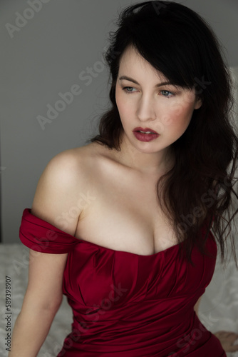 Portrait of a beautiful caucasian woman in her 30s wearing a red off the shoulder dress. She has dark brown hair with bangs. She is wearing red lipstick and her hair is messy. 