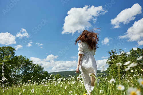 a woman with long red hair stands with her back to the camera in a light dress stands in a field of daisies against the blue sky and looks into the distance photo