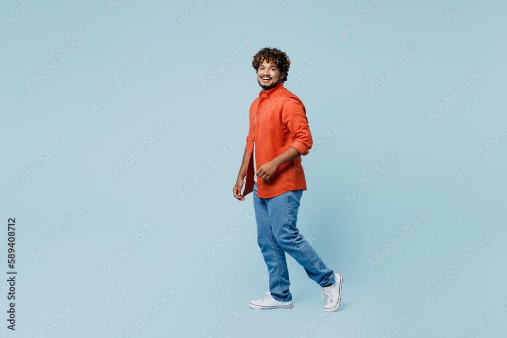Full body side profile view fun young Indian man wear orange red shirt white t-shirt walking going look camera isolated on plain pastel light blue cyan background studio portrait. Lifestyle concept.