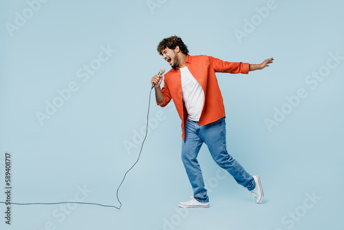 Foto Full body side profile view happy young Indian man wear orange red shirt white t-shirt sing song in microphone isolated on plain pastel light blue cyan background studio portrait