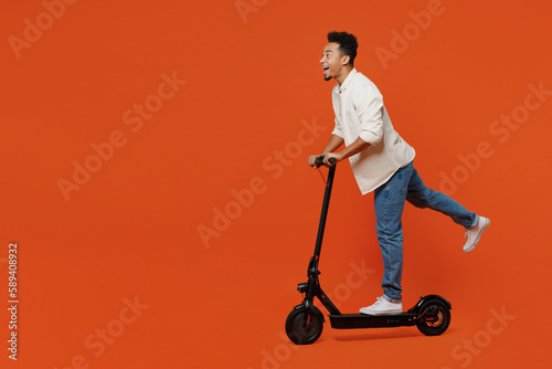 Full body side view overjoyed cool student young man of African American ethnicity wear light shirt casual clothes riding e-scooter isolated on orange red background studio portrait Lifestyle concept