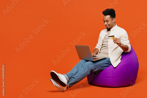 Full body young man of African American ethnicity in light casual clothes sit in bag chair use laptop pc computer hold credit bank card shopping online booking tour isolated on orange red background