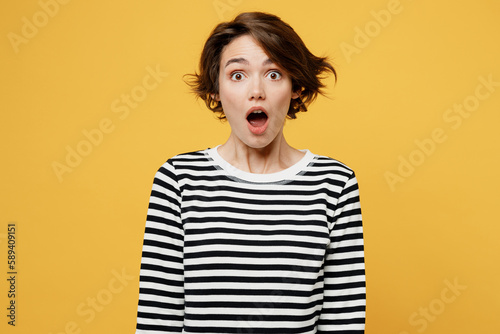 Young sad shocked indignant scared astonished stupefied woman wearing casual striped black and white shirt looking camera with opened mouth isolated on plain yellow color background studio portrait.