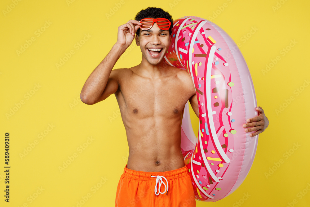 Handsome fun young sexy man wear orange shorts swimsuit take off sunglasses relax near hotel pool hold rubber donut ring isolated on plain yellow background. Summer vacation sea rest sun tan concept.