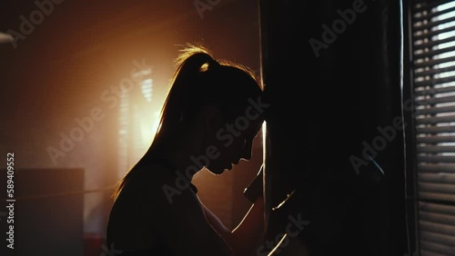 Silhouette of young tired exhausted sportswoman boxer loaning head and hands to punching bag in dark, side view. Sweaty girl is resting after hard training. Workout, box, professional sport concept. photo
