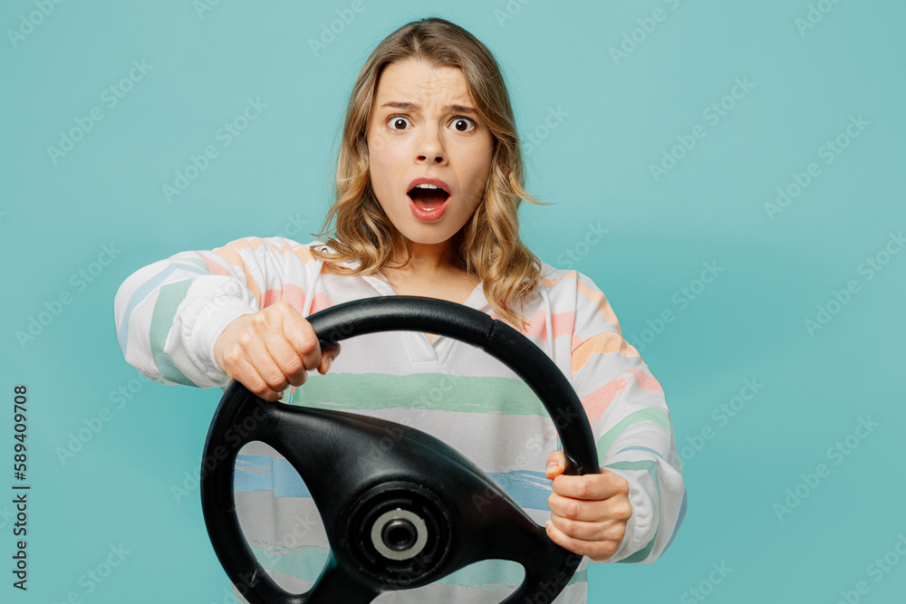 Young scared sad fearful woman wear striped hoody hold steering wheel driving car look camera with opened mouth isolated on plain pastel light blue cyan background studio portrait. Lifestyle concept. Young scared sad fearful woman wear striped hoody hold steering wheel driving car look camera with opened mouth isolated on plain pastel light blue cyan background studio portrait. Lifestyle concept.