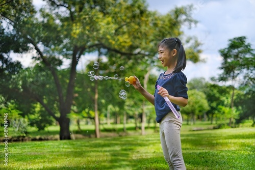 A cute young Asian girl is playing with bubbles using a bubble wands in a park on a bright sunny day with beautiful trees and green grass in the background.