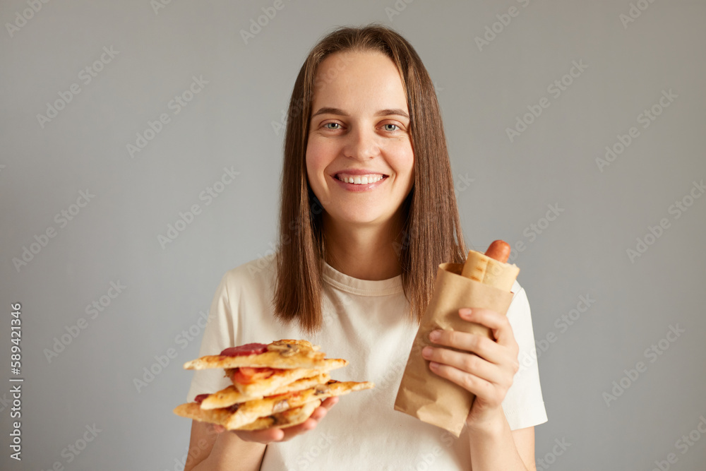 Portrait of young pretty girl holding hotdog and slices of pizza isolated on gray background, enjoying fast food, looking at camera with joyful cheerful expression.