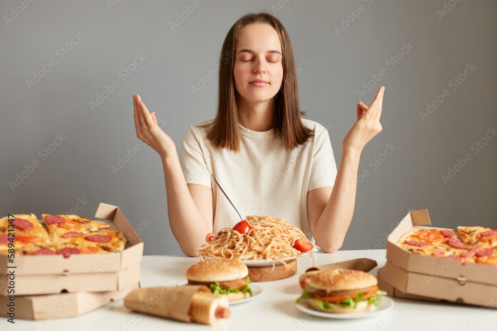 Indoor shot of calm relaxed woman with brown hair wearing white T-shirt sitting at table isolated over gray background, meditating before having dinner, saying thanks for tasty dish.