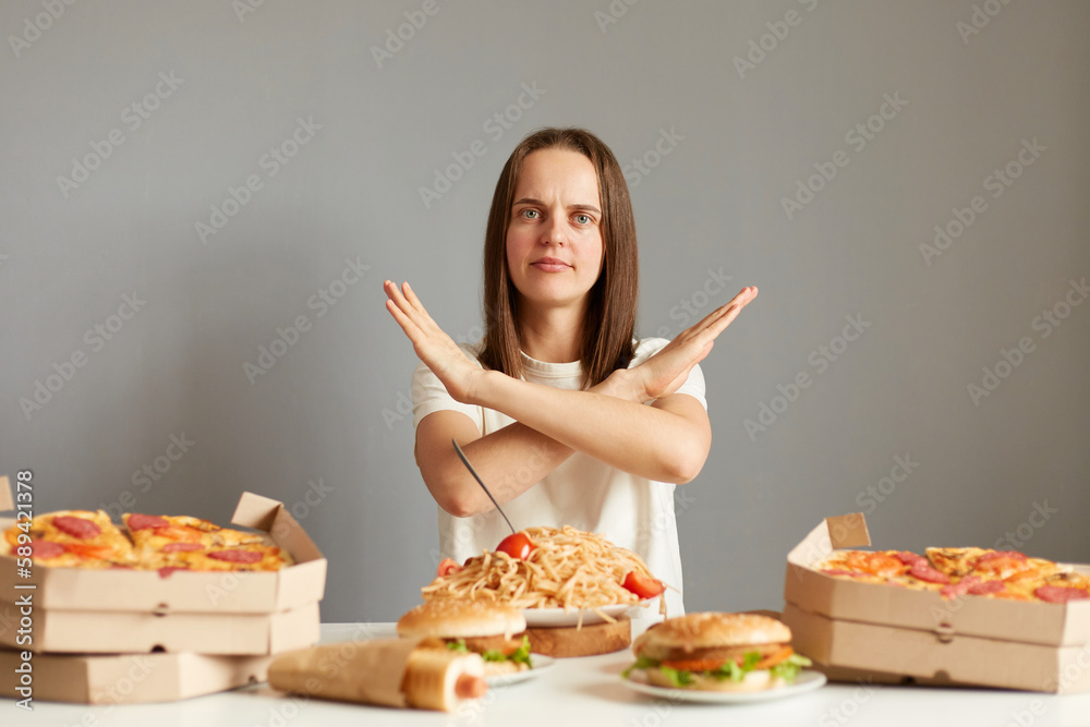 Portrait of attractive serious woman with brown hair wearing white T-shirt sitting at table isolated over gray background, showing no way gesture, keeps diet, gives up fast food. Portrait of attractive serious woman with brown hair wearing white T-shirt sitting at table isolated over gray background, showing no way gesture, keeps diet, gives up fast food.