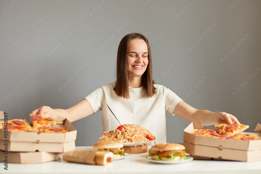Portrait of joyful charming woman with brown hair wearing white T-shirt sitting at table among different fast food, taking pizza from two boxes same time posing isolated over gray background Portrait of joyful charming woman with brown hair wearing white T-shirt sitting at table among different fast food, taking pizza from two boxes same time posing isolated over gray background
