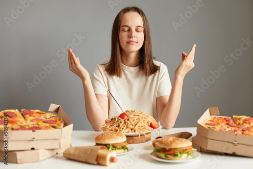 Indoor shot of calm relaxed woman with brown hair wearing white T-shirt sitting at table isolated over gray background  meditating before having dinner  saying thanks for tasty dish.