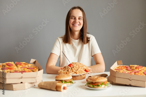 Indoor shot of woman with brown hair wearing white T-shirt sitting at table isolated over gray background, being surrounded tasty fast food, looking smiling at camera.