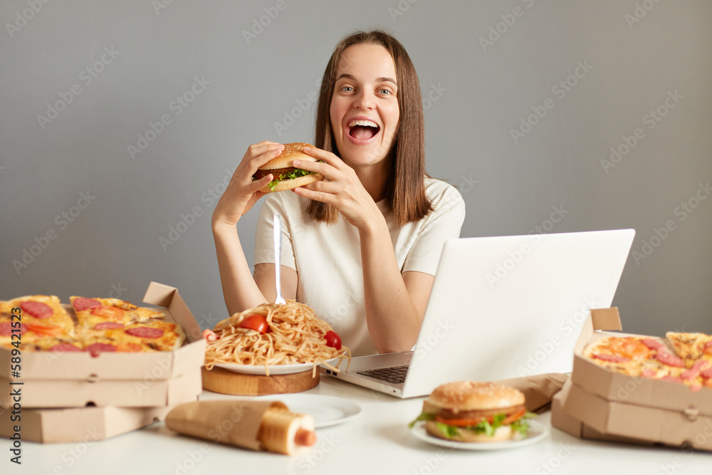 Portrait of overjoyed woman laptop sitting at table with computer and fast food, rejoicing ending diet, eating junk food, isolated over gray background. Portrait of overjoyed woman laptop sitting at table with computer and fast food, rejoicing ending diet, eating junk food, isolated over gray background.
