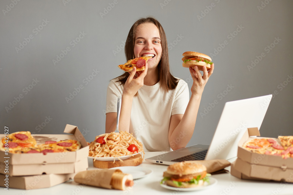 Portrait of satisfied woman sitting at table with laptop computer and different unhealthy food, isolated over gray background, biting pizza, enjoying delicious junk food dinner. Portrait of satisfied woman sitting at table with laptop computer and different unhealthy food, isolated over gray background, biting pizza, enjoying delicious junk food dinner.