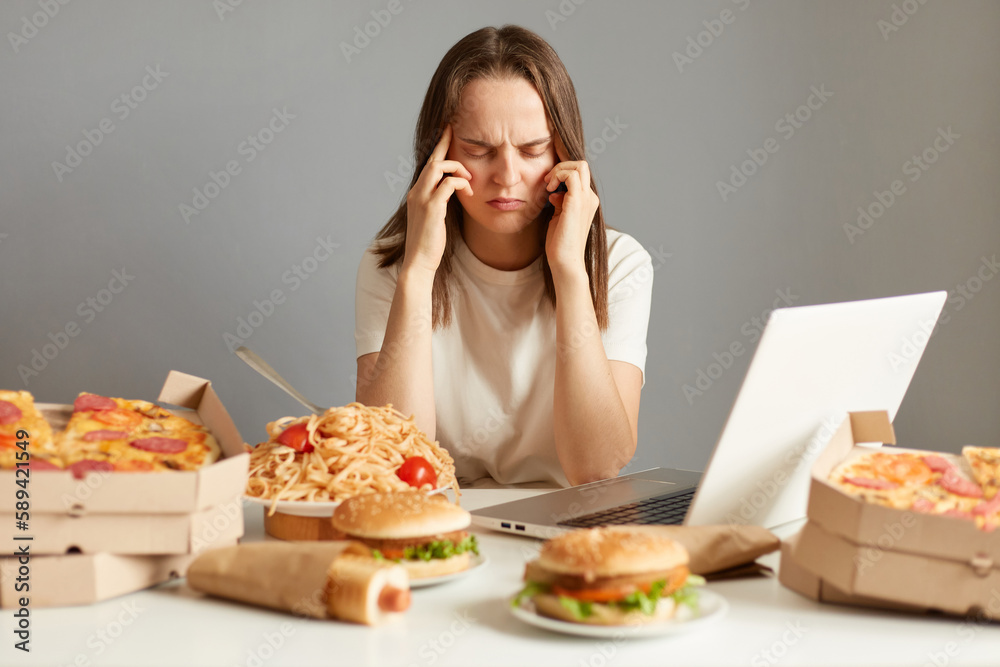 Indoor shot of sick unhealthy woman sitting in front of laptop among junk food, having terrible headache, being tire and exhausted, isolated over gray background. Indoor shot of sick unhealthy woman sitting in front of laptop among junk food, having terrible headache, being tire and exhausted, isolated over gray background.