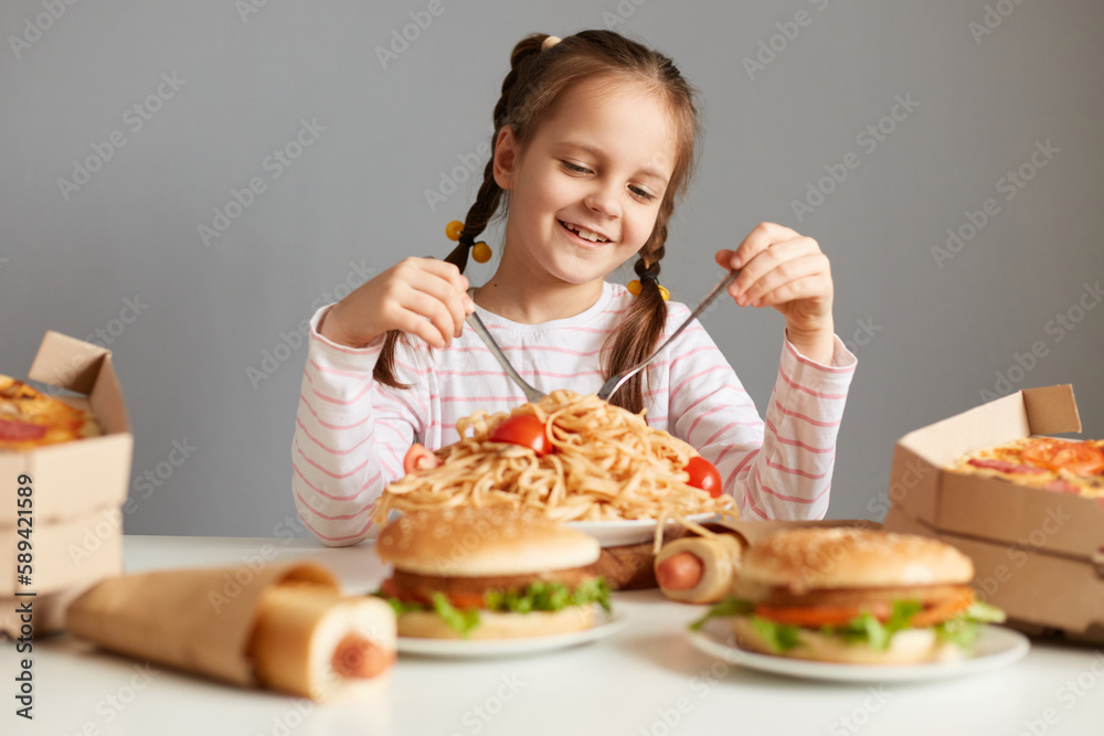 Portrait of satisfied little girl with pigtails sitting at table with big variety of junk food, eating pasta with satisfied expression, isolated over gray background