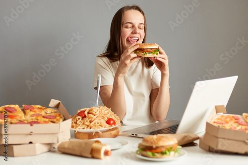 Portrait of hungry woman watching movie laptop computer  eating hamburger while resting  having cheat meal isolated over gray background  showing tongue out.