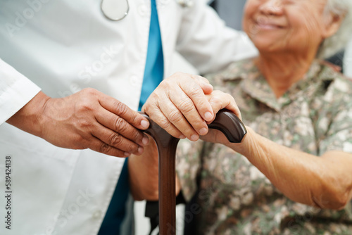 Doctor help Asian elderly disability woman patient holding walking stick in wrinkled hand at hospital. © manassanant