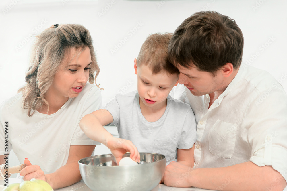 Young Caucasian family of three cooks lunch or festive cake in kitchen according to recipe. Happy family at table in bright kitchen.. Young Caucasian family of three cooks lunch or festive cake in kitchen according to recipe. Happy family at table in bright kitchen..
