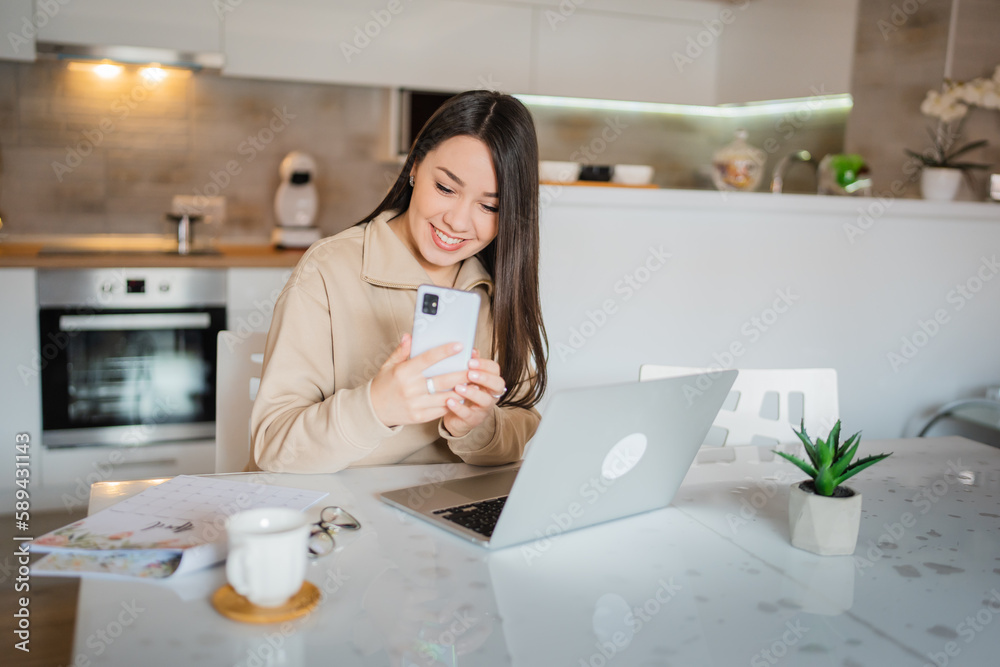 One young woman caucasian working at home in the kitchen and using a mobile phone, reading messages, modern lifestyle and business concept One young woman caucasian working at home in the kitchen and using a mobile phone, reading messages, modern lifestyle and business concept