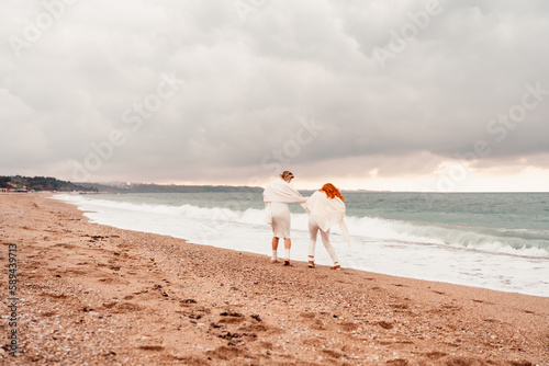 Women sea walk friendship spring. Two girlfriends  redhead and blonde  middle-aged walk along the sandy beach of the sea  dressed in white clothes. Against the backdrop of a cloudy sky and the winter