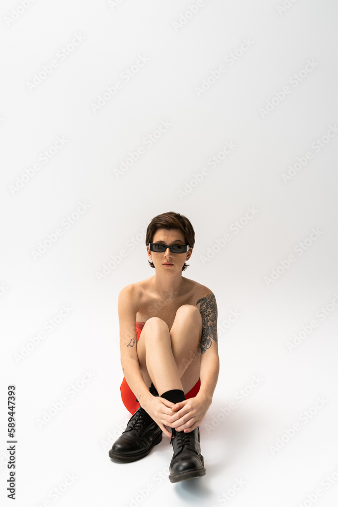 full length of young tattooed woman in black rough boots and trendy sunglasses sitting on grey background. full length of young tattooed woman in black rough boots and trendy sunglasses sitting on grey background.