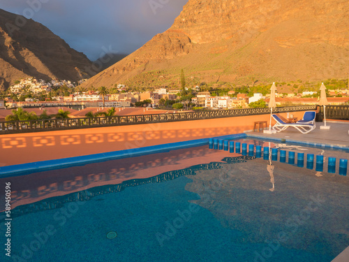 Aerial view of Valle Gran Rey with mountain cliffs, apartments and traditional houses in golden hour light. Seen over swimming pool on rooftop. La Gomera, Canary Islands, Spain. Holiday concept