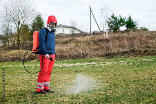Farmer spraying pesticide on lawn field wearing protective clothing. Treatment of grass from weeds and dandelion. Pest control. Insecticide sprayer with a proper protection. Gardening care season