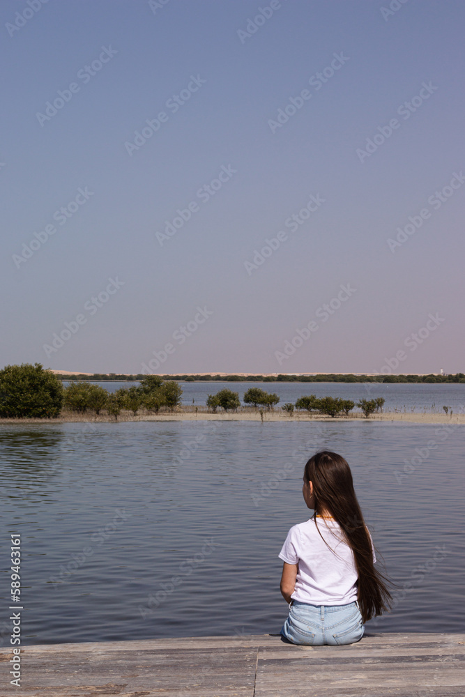A young girl sitting on a wooden gangway bridge looking at the sea water, waiting, good weather on a shore, blue skies, summer, mangroves