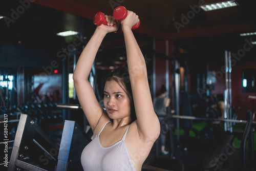 A young asian woman does standing tricep extensions with a pair of pink rubberized dumbbells. Working out triceps and arms at the gym.