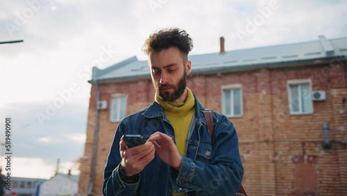 Horizontal shot of young confident man with stylish haircut using smartphone navigator, typing on screen keyboard and looking around. Portrait of lost attractive guy in city. Sunlight. Spring. Outside
