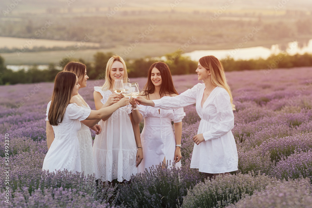 Five happy young girlfriends n the white dresses picnicking in the lavender field and toasting with glasses of white wine. 
