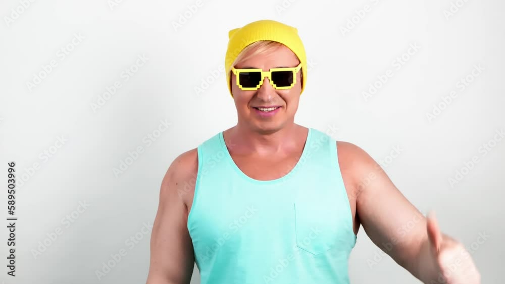 Young man showing thumb up, winner gesture in yellow beanie, blue tank top and sunglasses. Excited emotional guy celebrating success, victory. Body language. Studio shot isolated on white background. Young man showing thumb up, winner gesture in yellow beanie, blue tank top and sunglasses. Excited emotional guy celebrating success, victory. Body language. Studio shot isolated on white background.