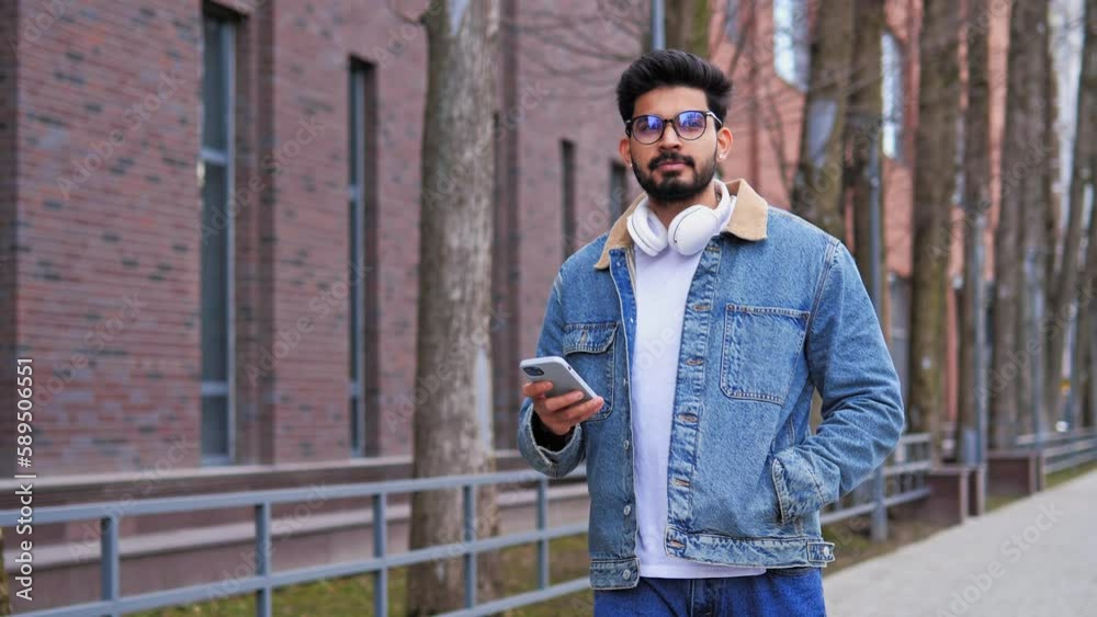 Good looking Latino man with headphones walks down the street, chatting on his mobile phone, typing a message while scrolling. Surfing social media, enjoying his free time and the holiday concept.