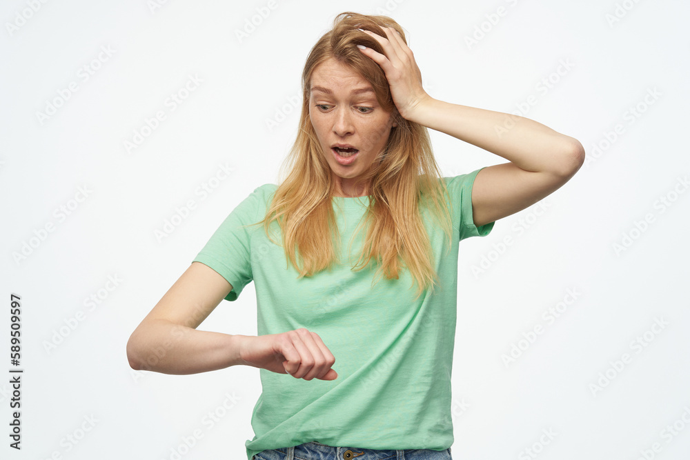 Indoor studio portrait of young ginger female with freckles looking at watches and touching her head isolated over background Indoor studio portrait of young ginger female with freckles looking at watches and touching her head isolated over background