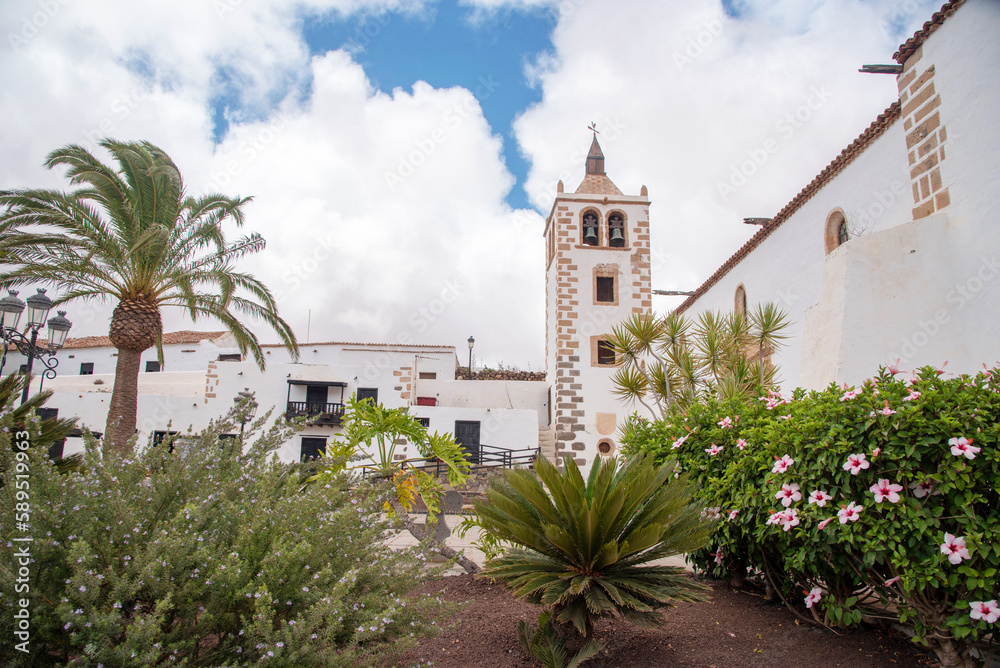 Vista panorámica de la plaza del pueblo de Betancuria en Fuerteventura, Islas Canarias con su impresionante iglesia blanca rodeada de mucha vegetación, cactus, palmeras y otras plantas típicas.