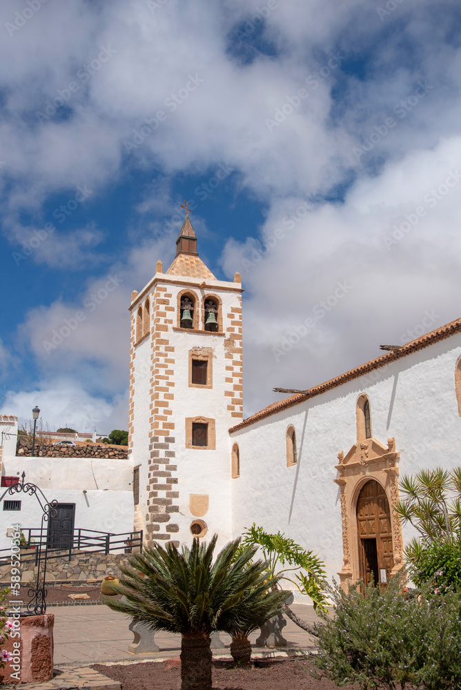 Plano de detalle del campanario de piedra blanca de la iglesia en el pueblo de Betancuria en Fuerteventura, Islas Canarias, rodeado de grandes palmeras y vegetación en un día soleado.