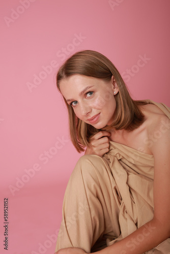 beautiful sexy blonde girl on pink background studio shot. A girl wrapped in a beige cloth sits on a pink background. beautiful girl model with make-up, stylish shooting