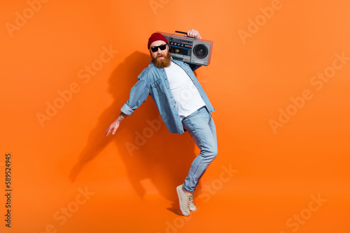 Full length photo of funky cheerful guy dressed denim jacket having fun listening boom box isolated orange color background